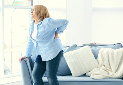 Una dona dempeus es toca la part baixa de l'esquena en senyal de dolor mentre es recolza en un sofà.