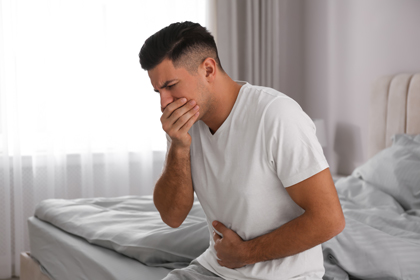Un home assegut en un llit fa un gest amb la mà a la boca que indica ganes de vomitar.